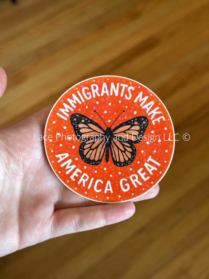 Puede incluir: Una pegatina redonda con el texto "IMMIGRANTS MAKE AMERICA GREAT" en letras blancas sobre un fondo naranja. En el centro hay una mariposa con alas negras y naranjas. La pegatina est&aacute; sujeta por una mano.