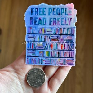 May include: A sticker with the text "FREE PEOPLE READ FREELY" above a watercolor illustration of a bookshelf. The sticker is held in a hand next to a quarter.
