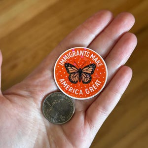 Puede incluir: Una pegatina redonda con el texto "IMMIGRANTS MAKE AMERICA GREAT" en blanco sobre un fondo naranja. La pegatina presenta una ilustraci&oacute;n de mariposa. Una moneda de 25 centavos estadounidense est&aacute; al lado de la pegatina.