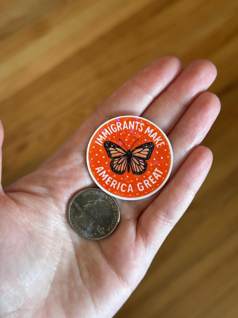 Puede incluir: Una pegatina redonda con el texto "IMMIGRANTS MAKE AMERICA GREAT" en blanco sobre un fondo naranja. La pegatina presenta una ilustraci&oacute;n de mariposa. Una moneda de 25 centavos estadounidense est&aacute; al lado de la pegatina.