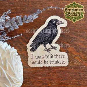 May include: A die-cut sticker featuring a black crow illustration with a wide-eyed expression. Below the bird, the text reads, I was told there would be trinkets, in a black gothic-style font on a cream-colored background with a die-cut edge.