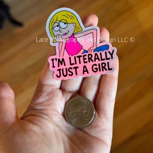 May include: A sticker featuring a cartoon girl with blonde hair and glasses, wearing a pink top and blue pants. The sticker has the text "I'M LITERALLY JUST A GIRL" in pink. A quarter is next to the sticker.