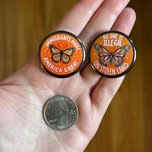 May include: Two round, orange and black buttons with butterfly illustrations and text. One reads "IMMIGRANTS MAKE AMERICA GREAT," the other says "NO ONE IS ILLEGAL ON STOLEN LAND." A quarter coin is also visible.