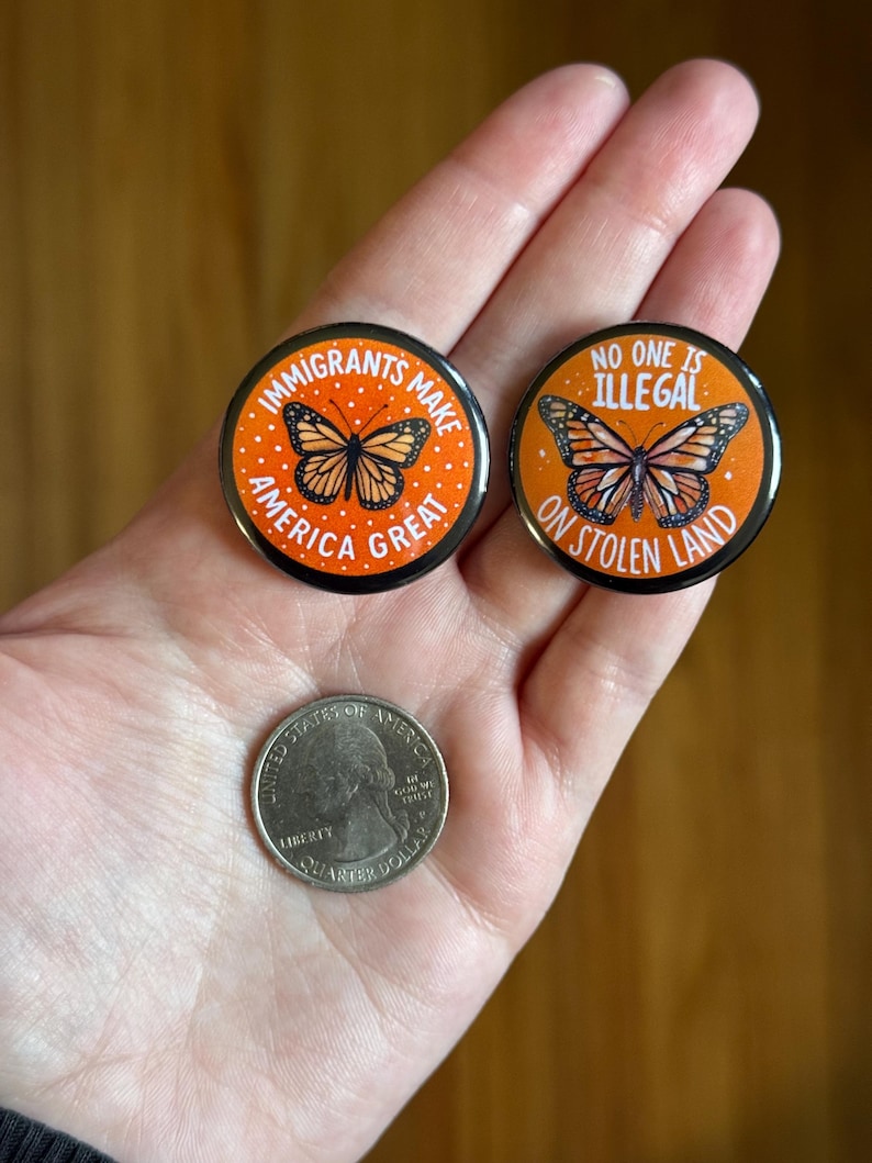 May include: Two round, orange and black buttons with butterfly illustrations and text. One reads "IMMIGRANTS MAKE AMERICA GREAT," the other says "NO ONE IS ILLEGAL ON STOLEN LAND." A quarter coin is also visible.