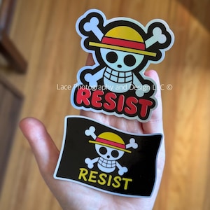 May include: Two stickers featuring a skull and crossbones design with a yellow hat and the word "RESIST". One sticker is a skull and crossbones with the word "RESIST" below it. The other sticker is a skull and crossbones with the word "RESIST" below it.