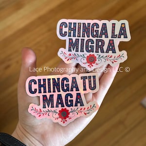 May include: Two holographic stickers with the phrases "CHINGA LA MIGRA" and "CHINGA TU MAGA" in navy blue, with floral accents in red and white. The stickers are held in a hand against a wooden background.