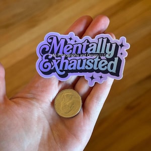 May include: A sticker with the words "Mentally Exhausted" in a purple and blue gradient design. The sticker has a decorative font and star accents. A gold coin is next to the sticker.