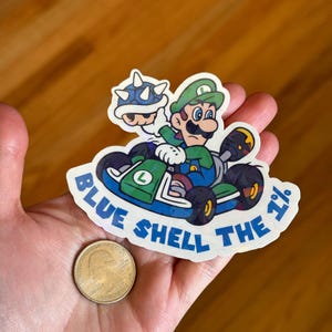 May include: A sticker featuring a green Mario character driving a blue kart with a blue shell in the back. The text "Blue Shell the 1%" is written around the sticker.