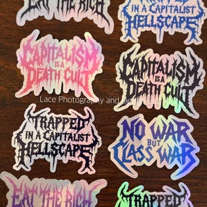 May include: A collection of holographic stickers with bold, gothic-style text. Phrases include "Eat the Rich," "Trapped in a Capitalist Hellscape," and "Capitalism is a Death Cult." The stickers have a dark, edgy aesthetic.