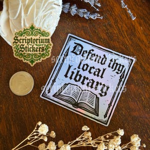 May include: A square sticker with the text "Defend thy local library" and an illustration of an open book. The sticker has a gradient background of blue and pink. Also visible is a sticker with the text "Scriptorium Stickers".