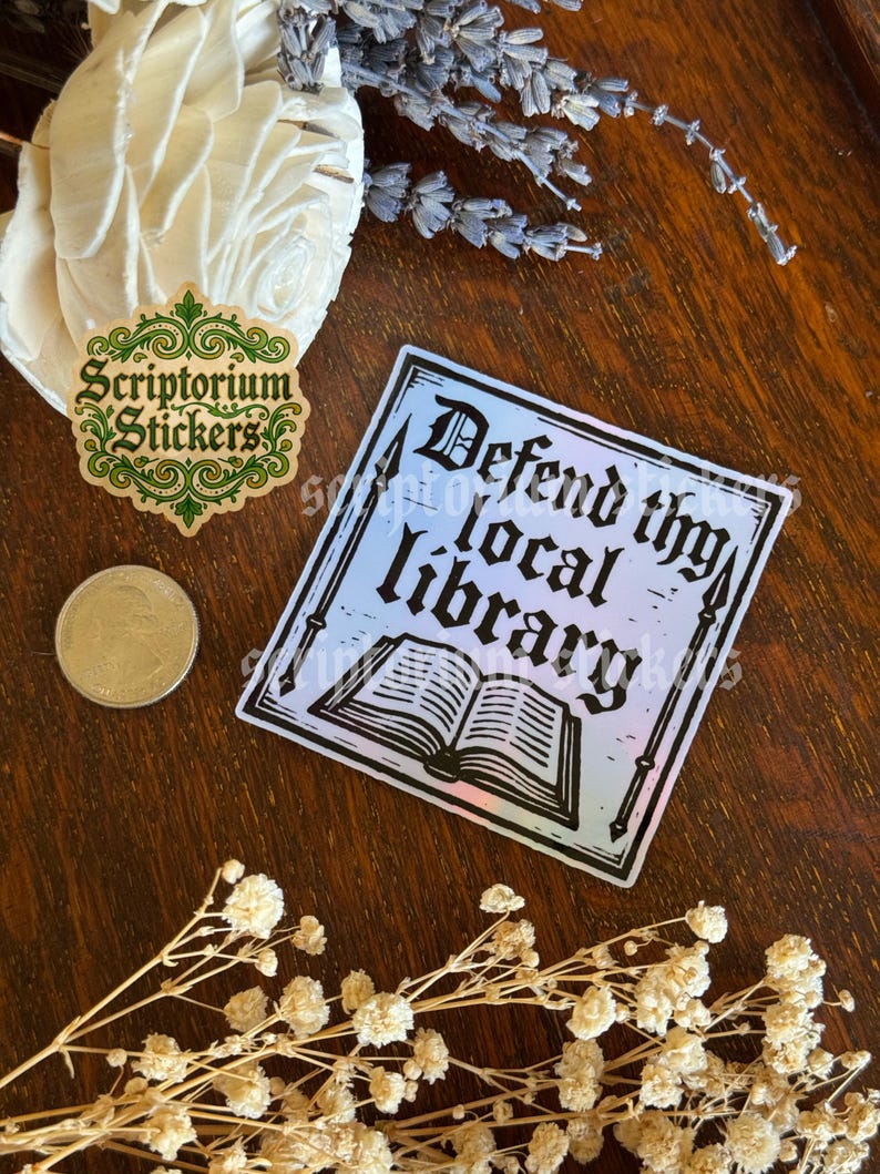 May include: A square sticker with the text "Defend thy local library" and an illustration of an open book. The sticker has a gradient background of blue and pink. Also visible is a sticker with the text "Scriptorium Stickers".