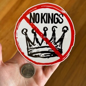 May include: A white sticker with a red circle and a red line through a black crown. The words "NO KINGS" are above the crown in black. A quarter is next to the sticker.