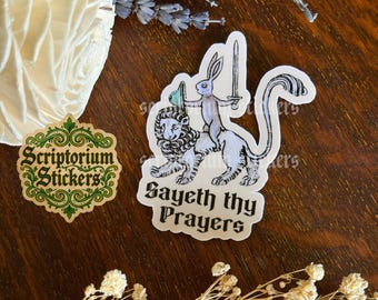 Pode incluir: Duas etiquetas adesivas numa superfície de madeira. Uma etiqueta apresenta um leão montado por um coelho com uma espada, com o texto "Sayeth thy Prayers". A outra etiqueta tem o texto "Scriptorium Stickers" numa moldura decorativa.