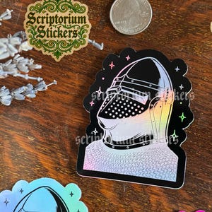May include: A collection of stickers featuring a knight's helmet, a logo with the text "Scriptorium Stickers", and a quarter. The knight's helmet sticker has a black background with a holographic design. The logo is green and gold.