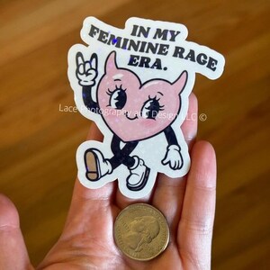 以下が含まれることがあります： 「IN MY FEMININE RAGE ERA」の文字が入った、手足のついた漫画のハートのステッカー。ロックオンのジェスチャーをしています。ステッカーの隣にはコインがあります。