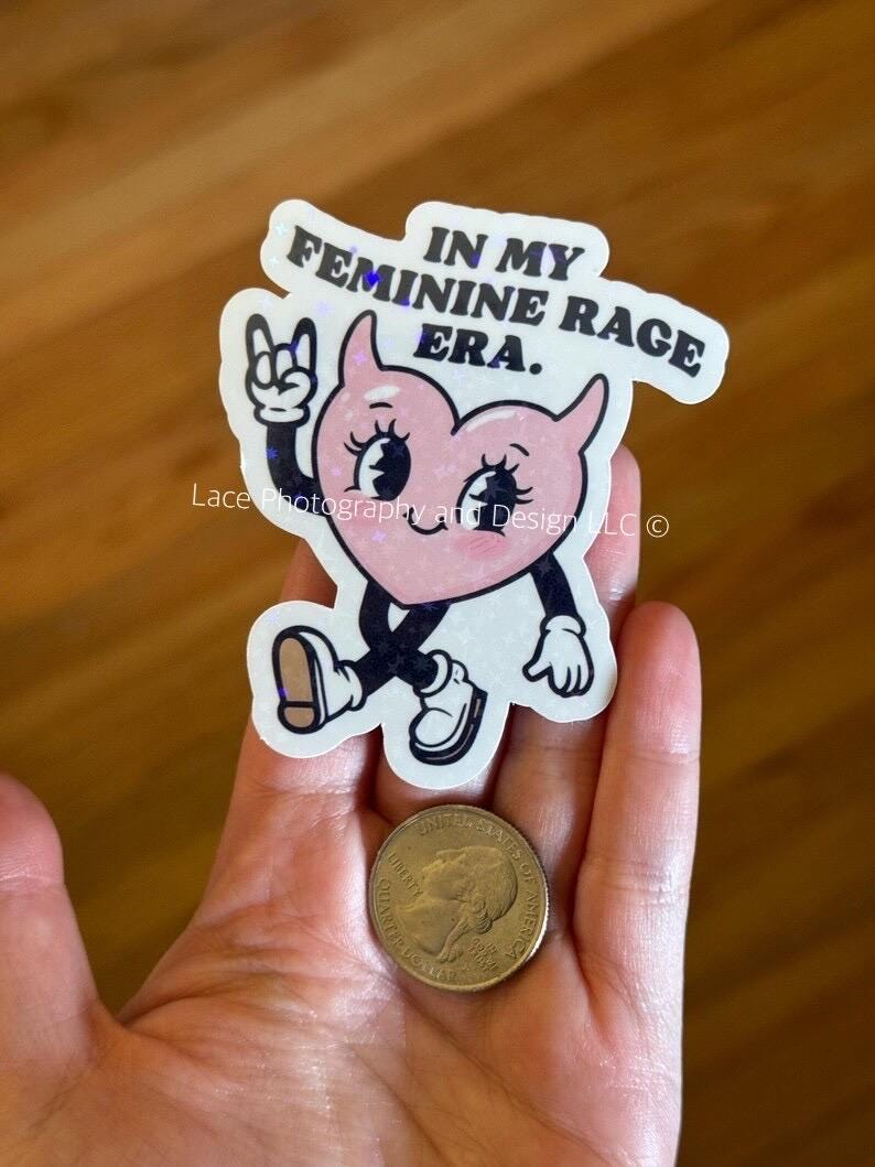 以下が含まれることがあります： 「IN MY FEMININE RAGE ERA」の文字が入った、手足のついた漫画のハートのステッカー。ロックオンのジェスチャーをしています。ステッカーの隣にはコインがあります。