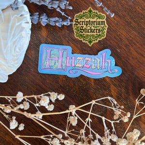Puede incluir: Una pegatina con la palabra "Huzzah" en una fuente caprichosa, delineada en rosa y azul, con un fondo azul claro. Tambi&eacute;n es visible una segunda pegatina con el texto "Scriptorium Stickers". Flores secas y lavanda dispuestas alrededor de las pegatinas.