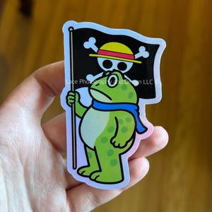 May include: A green frog sticker with a blue scarf holding a black flag with a white skull and crossbones and a yellow hat. The sticker has a holographic finish and the text "Lace Photography and Design LLC".