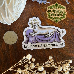 K&ouml;nnte beinhalten: Ein Aufkleber mit einer Katzenillustration auf einer lila Oberfl&auml;che, mit dem Text "Let them eat Temptations!". Der Aufkleber enth&auml;lt auch den Text "Scriptorium Stickers". Eine M&uuml;nze, Trockenblumen und ein dekoratives wei&szlig;es Objekt sind ebenfalls sichtbar.