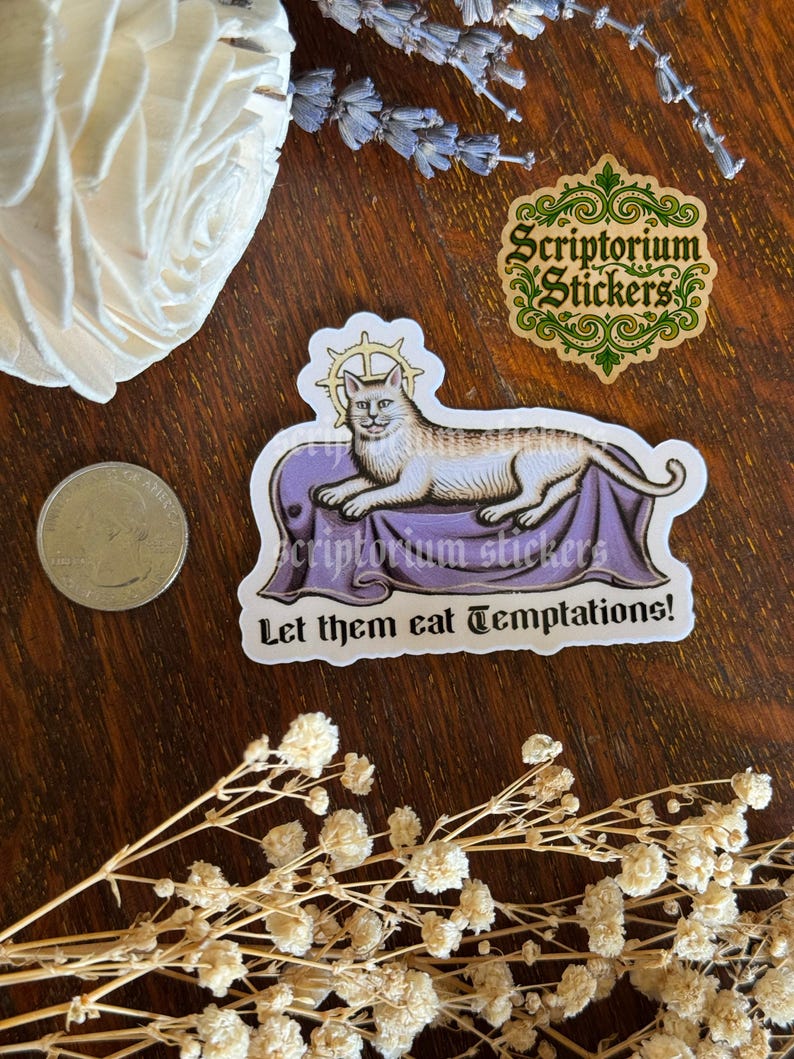 K&ouml;nnte beinhalten: Ein Aufkleber mit einer Katzenillustration auf einer lila Oberfl&auml;che, mit dem Text "Let them eat Temptations!". Der Aufkleber enth&auml;lt auch den Text "Scriptorium Stickers". Eine M&uuml;nze, Trockenblumen und ein dekoratives wei&szlig;es Objekt sind ebenfalls sichtbar.