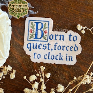 May include: A sticker with the text "Born to quest, forced to clock in" in a blue, medieval-style font. The sticker has a blue border with a large blue letter "B" and red floral accents. The background is a dark wood surface.