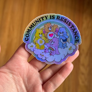 May include: A holographic sticker featuring a group of cartoon bears on a cloud with a rainbow. The sticker has the text "COMMUNITY IS RESISTANCE" in a semi-circle around the bears. The bears are in various colors, including yellow, orange, pink, and blue.