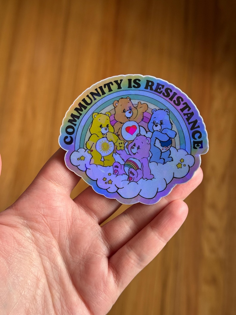 May include: A holographic sticker featuring a group of cartoon bears on a cloud with a rainbow. The sticker has the text "COMMUNITY IS RESISTANCE" in a semi-circle around the bears. The bears are in various colors, including yellow, orange, pink, and blue.
