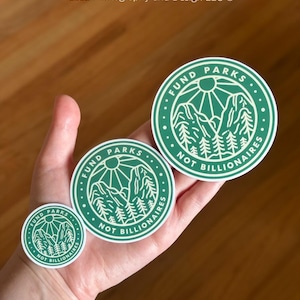 May include: Three green and white round stickers with the text "Fund Parks Not Billionaires" and a graphic of mountains and trees. The stickers are different sizes.