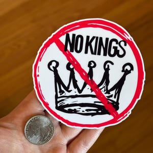 May include: A round sticker with the words "NO KINGS" in black, crossed out with a red line. A black crown is below the text. The sticker is white with a red border. A quarter is next to the sticker.