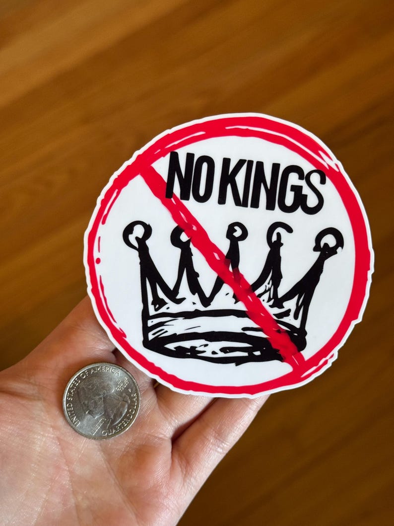 May include: A round sticker with the words "NO KINGS" in black, crossed out with a red line. A black crown is below the text. The sticker is white with a red border. A quarter is next to the sticker.