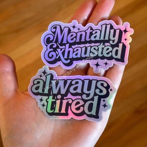 May include: Two holographic stickers with black outlines and text. The top sticker reads "Mentally Exhausted" in a stylized font. The bottom sticker says "always tired". Both stickers have star accents and a rainbow sheen.