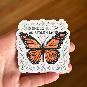 May include: A sticker featuring a pixelated monarch butterfly with orange and black wings. The sticker has the text "NO ONE IS ILLEGAL ON STOLEN LAND" in black. The sticker is surrounded by small floral designs.