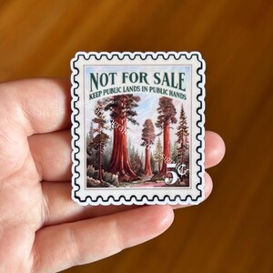 May include: A sticker in the shape of a postage stamp featuring a forest scene with large redwood trees. The text "NOT FOR SALE" and "KEEP PUBLIC LANDS IN PUBLIC HANDS" are printed on the sticker. The sticker is held in a hand.