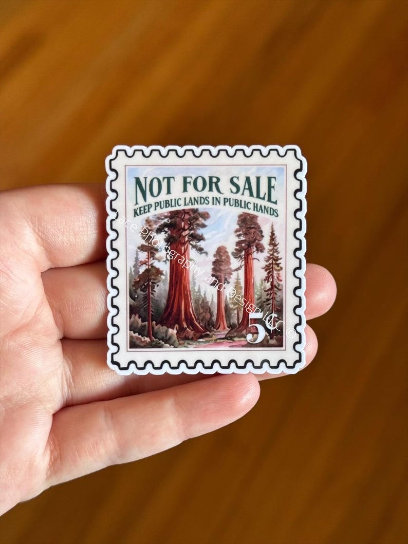 May include: A sticker in the shape of a postage stamp featuring a forest scene with large redwood trees. The text "NOT FOR SALE" and "KEEP PUBLIC LANDS IN PUBLIC HANDS" are printed on the sticker. The sticker is held in a hand.