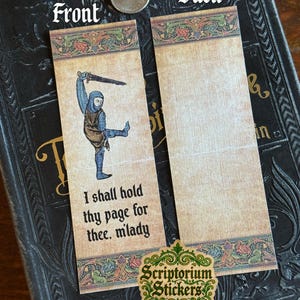 May include: Two medieval-themed bookmarks. The front bookmark features a knight illustration with the text "I shall hold thy page for thee, milady." The back bookmark is blank. The bookmarks are on a dark book.