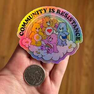 May include: A holographic sticker featuring a rainbow and several cartoon bears on a cloud. The sticker has the text "COMMUNITY IS RESISTANCE" in a rainbow arc. A quarter is next to the sticker for size comparison.