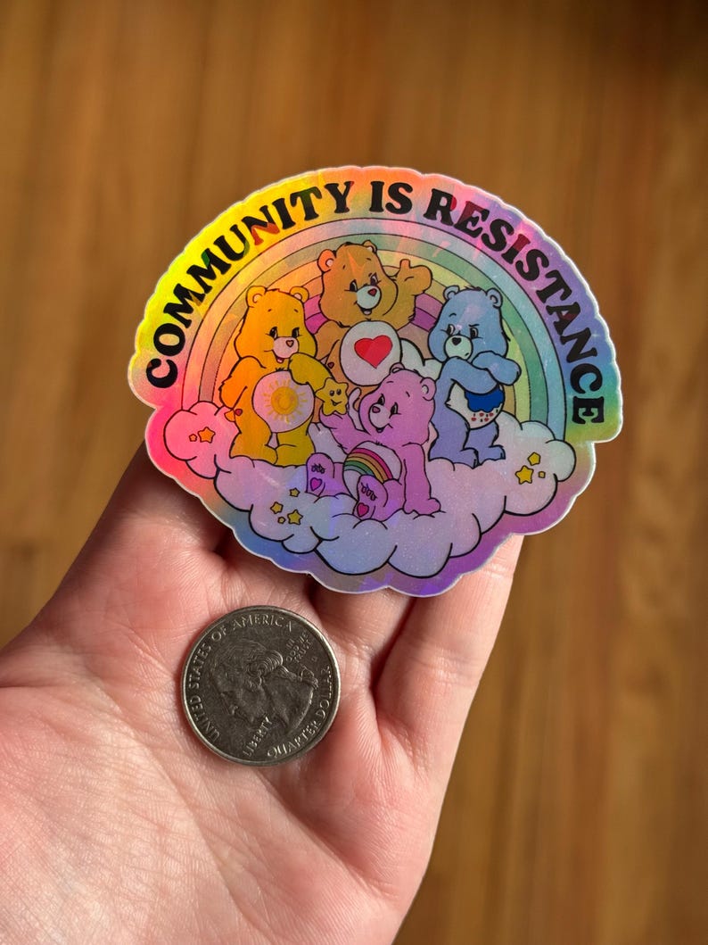 May include: A holographic sticker featuring a rainbow and several cartoon bears on a cloud. The sticker has the text "COMMUNITY IS RESISTANCE" in a rainbow arc. A quarter is next to the sticker for size comparison.