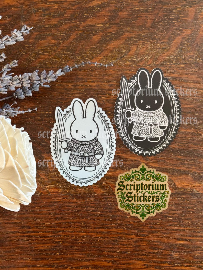 May include: Two oval stickers featuring a white rabbit character wearing chainmail armor and holding a sword, framed by a decorative border. One sticker has a white background and the other a black background. A third sticker displays the text Scriptorium Stickers.