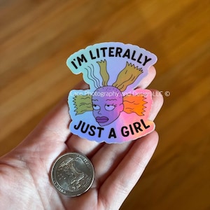 May include: Holographic sticker featuring a cartoon character with the text "I'm Literally Just a Girl." The character has purple skin, blonde and orange hair, and a stern expression. A quarter is next to the sticker.