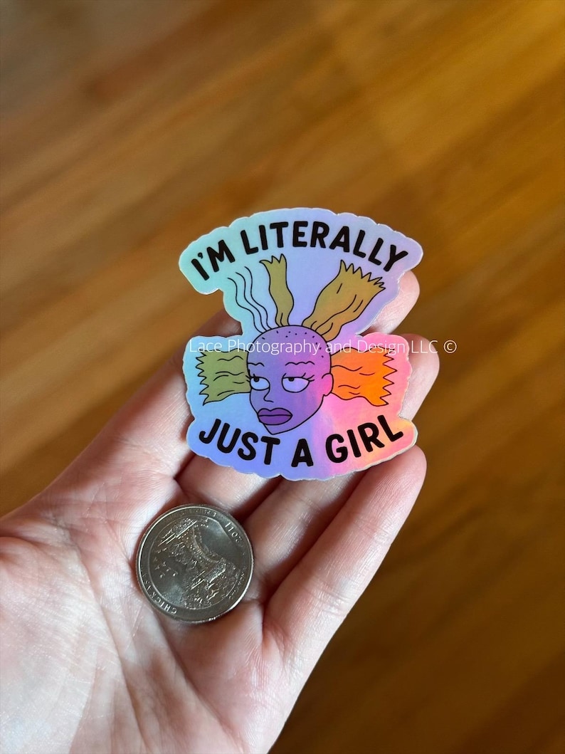 May include: Holographic sticker featuring a cartoon character with the text "I'm Literally Just a Girl." The character has purple skin, blonde and orange hair, and a stern expression. A quarter is next to the sticker.