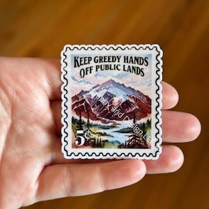May include: A sticker resembling a vintage postage stamp, featuring a mountain landscape with a lake and trees. The text "KEEP GREEDY HANDS OFF PUBLIC LANDS" is displayed above the mountain scene. The stamp also includes a "5&cent;" value.