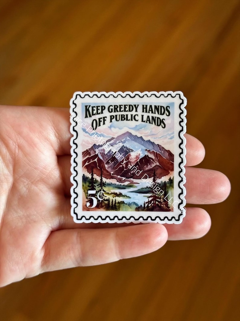 May include: A sticker resembling a vintage postage stamp, featuring a mountain landscape with a lake and trees. The text "KEEP GREEDY HANDS OFF PUBLIC LANDS" is displayed above the mountain scene. The stamp also includes a "5&cent;" value.