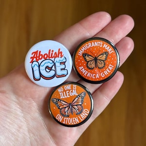 May include: Three round buttons with political statements. One button says "Abolish ICE" in red and blue. The other two buttons have orange backgrounds with butterfly illustrations and the phrases "Immigrants Make America Great" and "No One Is Illegal On Stolen Land."
