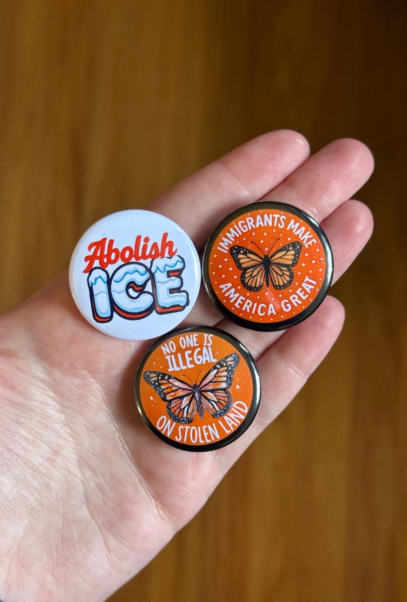 May include: Three round buttons with political statements. One button says "Abolish ICE" in red and blue. The other two buttons have orange backgrounds with butterfly illustrations and the phrases "Immigrants Make America Great" and "No One Is Illegal On Stolen Land."