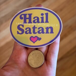 Pode incluir: Um adesivo oval com as palavras "Hail Satan" em escrita roxa sobre um fundo amarelo, com uma borda roxa e um cora&ccedil;&atilde;o rosa. Uma moeda de vinte e cinco centavos americana est&aacute; ao lado do adesivo.