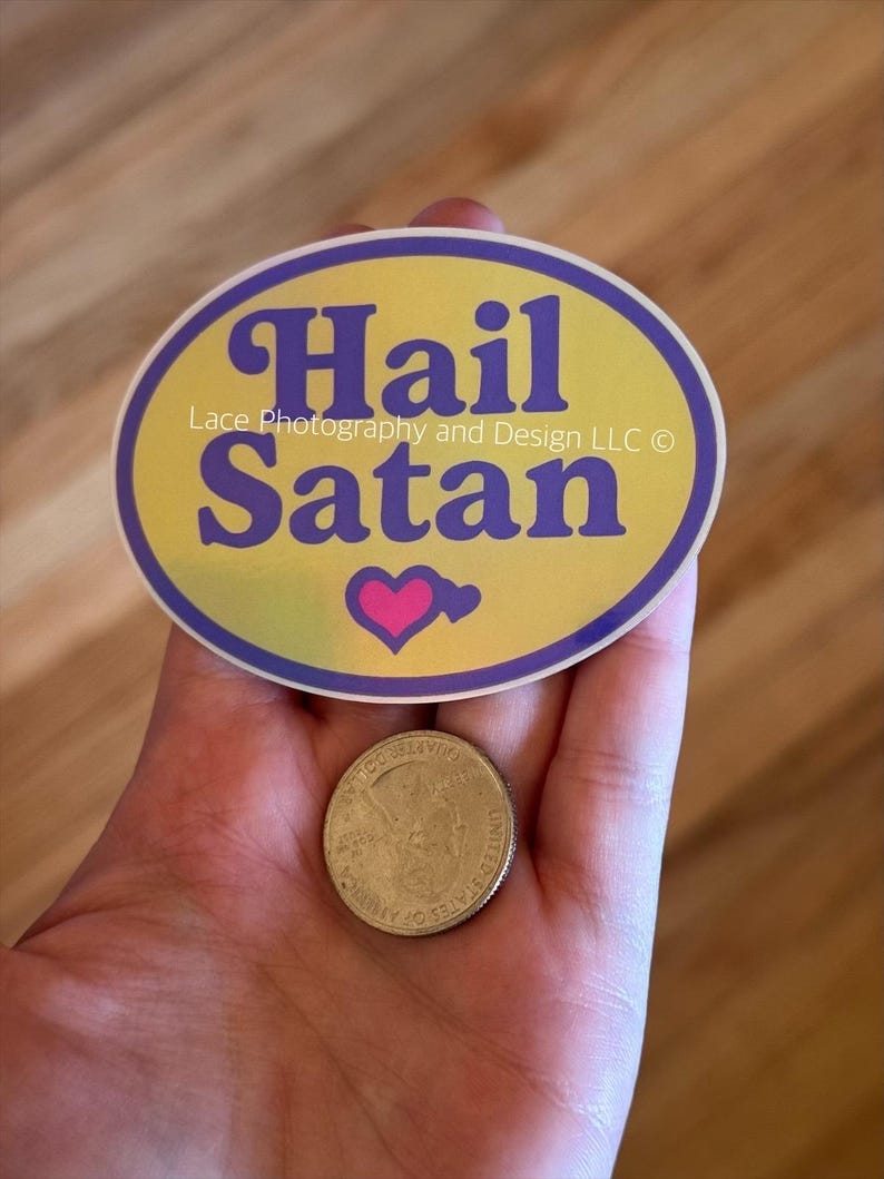 Pode incluir: Um adesivo oval com as palavras "Hail Satan" em escrita roxa sobre um fundo amarelo, com uma borda roxa e um cora&ccedil;&atilde;o rosa. Uma moeda de vinte e cinco centavos americana est&aacute; ao lado do adesivo.