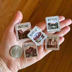 May include: A collection of miniature postage stamps with mountain and forest landscape designs. The stamps feature text such as "Protect Our Public Lands" and "Resist". A quarter is also visible.