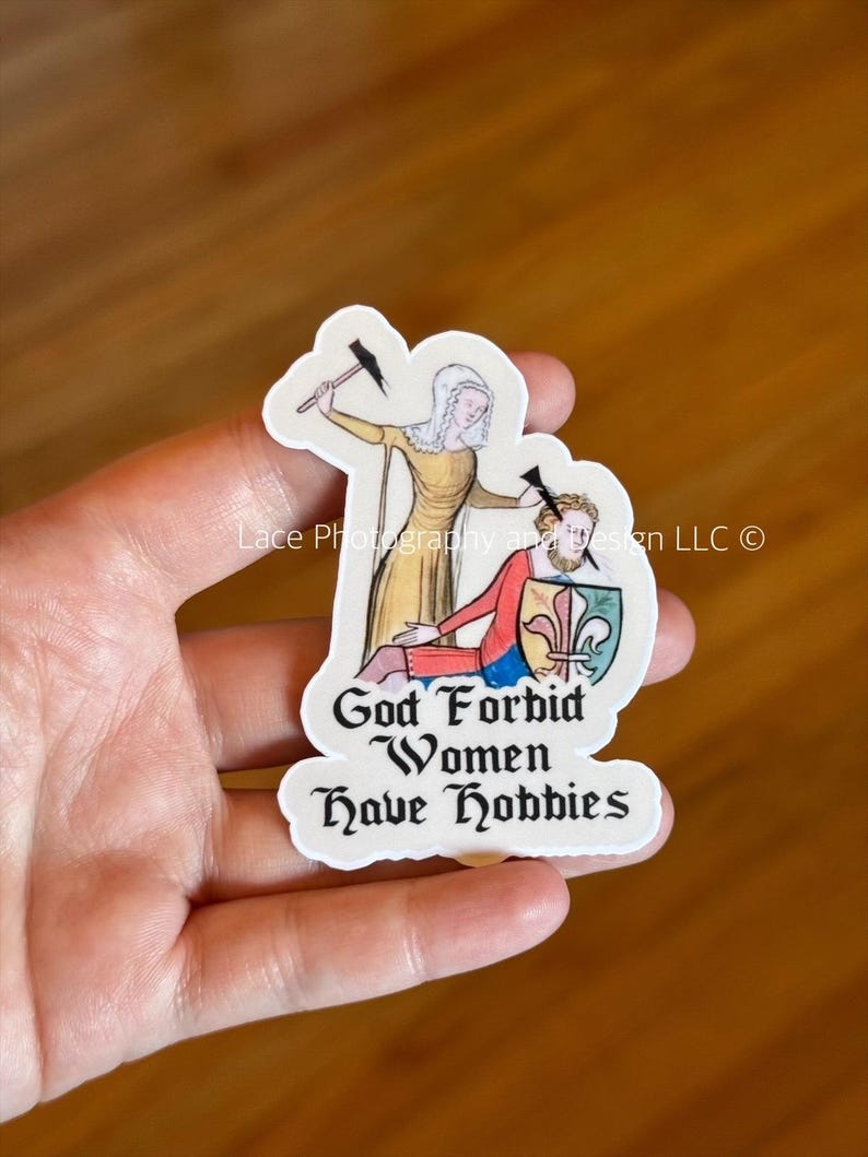 May include: A sticker featuring a medieval illustration of a woman in a yellow dress with a hammer, and a man in red and blue. The text "God Forbid Women Have Hobbies" is below the figures. The sticker has a white border.