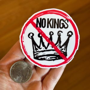 May include: A white sticker with a red border and the words "NO KINGS" in black, crossed out with a red line. Below the text is a black crown, also crossed out. A quarter is next to the sticker.