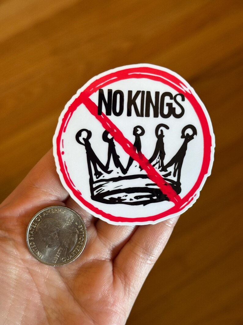May include: A white sticker with a red border and the words "NO KINGS" in black, crossed out with a red line. Below the text is a black crown, also crossed out. A quarter is next to the sticker.